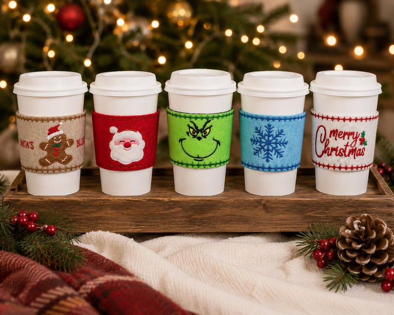 Op de afbeelding: Vijf witte wegwerp koffiebekers met vilten hoezen. De hoezen zijn voorzien van een peperkoekmannetje, een kerstmangezicht, een groen karaktergezicht, een blauwe sneeuwvlok en de tekst Merry Christmas met een hulsttakje.