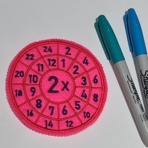 May include: A pink felt circle with a 2x multiplication table printed on it. The numbers 1 through 12 are printed around the outside of the circle, and the numbers 2 through 24 are printed in the center of the circle.