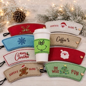 Op de afbeelding: Een collectie vilten koffiebekerhoezen met diverse vakantiemotieven, waaronder een groen Grinch-gezicht, de Kerstman, rendieren, sneeuwvlokken, peperkoekmannetjes en kerstbomen met teksten zoals Santa's Blend en Oh What Fun.