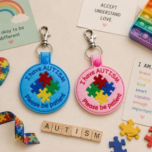 May include: Two circular keychains with silver lobster clasps, one light blue and one pink. Both feature embroidered text "I have AUTISM Please be patient" and a four-piece colorful puzzle piece graphic in red, green, yellow, and blue.