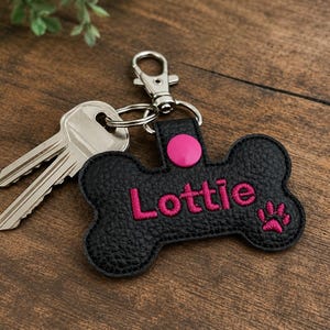 May include: A black bone-shaped keychain made of textured material featuring the name Lottie embroidered in bright pink thread, accompanied by a small pink paw print icon, a pink snap button, and a silver metal lobster clasp holding two silver keys.