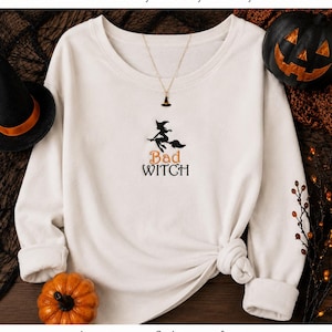 Könnte beinhalten: Ein weißes Langarm-Sweatshirt mit einer schwarzen Stickerei einer Hexensilhouette auf einem Besen über dem orange-schwarzen Text "Bad Witch". Erhältlich in vier Größen: 100mm, 130mm, 160mm, 200mm, mit den Dateiformaten pes, hus, vip, xxx, vp3, dst, exp, jef, pec und sew.