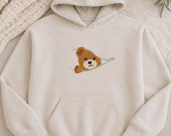 May include: A beige hooded sweatshirt featuring a central embroidered graphic of a brown teddy bear head peeking out from behind a silver zipper design. The hoodie includes a front kangaroo pocket, long sleeves, and a matching drawstring hood.