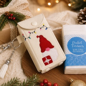 May include: A cream-colored rectangular tissue holder featuring an embroidered gnome wearing a red hat and holding a red gift, accented with colorful string lights. It includes a silver metal keychain with a white tassel and a snap closure.