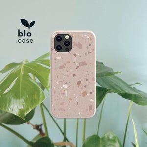 May include: A phone case with a light beige base featuring a speckled terrazzo pattern of small pink, brown, and white geometric shapes. The case has a cutout for a triple-lens camera system. The text bio case is printed in black with a leaf icon.