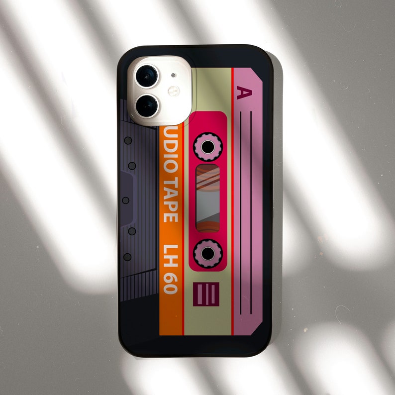 May include: A black phone case with a retro cassette tape design. The cassette tape is orange, pink, and yellow with the text "AUDIO TAPE LH 60" and "A" on it.