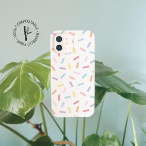 May include: A white phone case featuring a scattered pattern of small, rectangular confetti shapes in pastel pink, yellow, blue, and purple. A circular logo in the upper left corner contains the text 100% COMPOSTABLE BAMBOO FIBER.