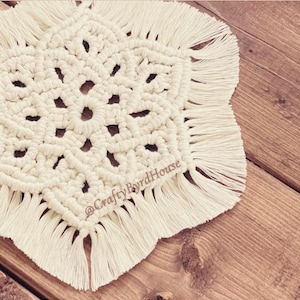 May include: Cream-colored macrame coaster with a geometric design and fringe detail. The coaster is handmade and features the text "@CraftyByrdHouse". This decorative item is perfect for adding a touch of bohemian style to any home.