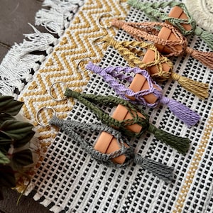 May include: Six macrame plant hangers with terracotta pots and keychains. The hangers are in various colors, including green, purple, and gray. Each hanger has a tassel at the bottom.