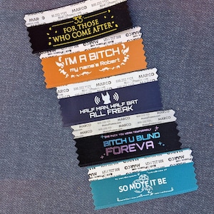 May include: A collection of fabric labels with printed text. The labels are in black, orange, navy blue, and teal. Text includes phrases such as "FOR THOSE WHO COME AFTER" and "I'M A BITCH my name's Robert".