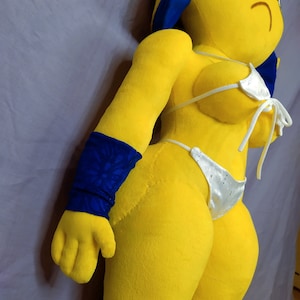 Puede incluir: Un juguete de peluche amarillo con una diadema azul, que lleva un top y una parte inferior de bikini blancos. El juguete tiene un brazalete de brazo con estampado azul y negro.