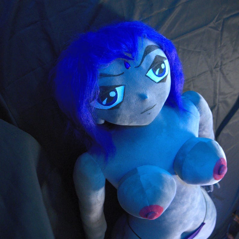 May include: A plush doll of a cartoon character with blue skin, hair, and eyes. The doll has a stern expression and is wearing a purple bikini. The doll's breasts have pink nipples. The doll is lying on a dark surface.