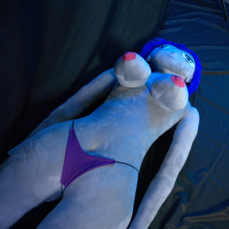 May include: A plush doll in a reclining pose, featuring a light blue body, blue hair, and a purple thong. The doll has large breasts with pink nipples. The background is a dark, possibly black, surface.