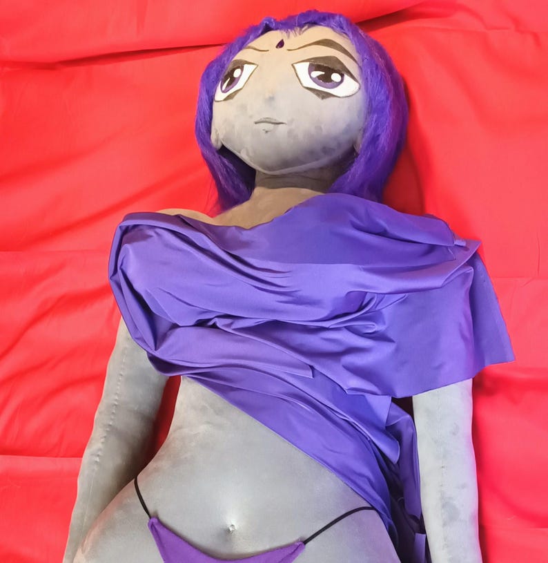 May include: A plush doll of a character with gray skin, purple hair, and a purple outfit. The doll is lying on a red surface. The doll's outfit includes a draped top and a bikini bottom.