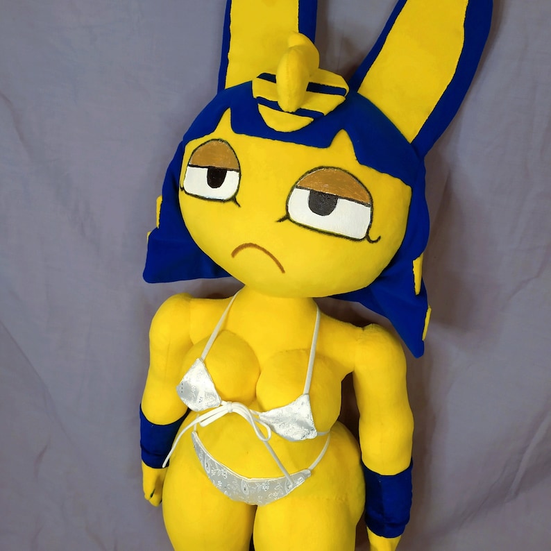 Puede incluir: Un juguete de peluche amarillo con pelo y orejas azules, que lleva un top y una parte inferior de bikini blancos. El juguete tiene una expresi&oacute;n triste en su rostro.