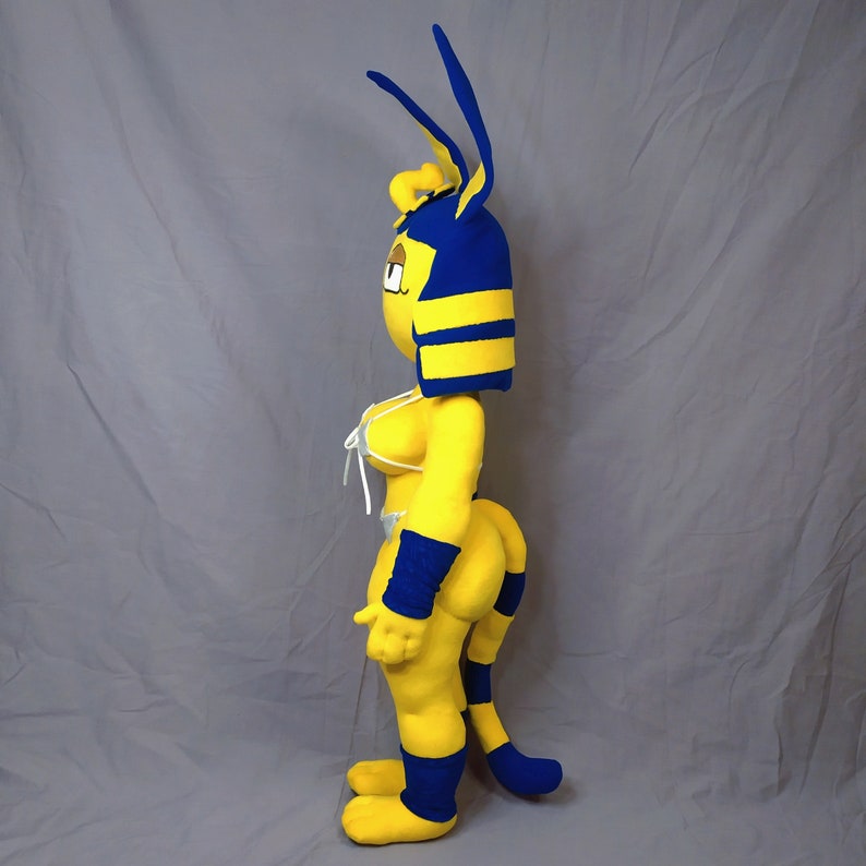 Puede incluir: Un juguete de peluche amarillo y azul con cara de gato y cola larga. El juguete lleva un tocado azul y amarillo y un top de bikini blanco.