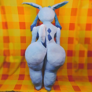 May include: A light blue plush doll with large, rounded features, including a prominent rear and legs. The doll has long ears and a tail, with darker blue accents. It is set against a yellow and orange checkered background.