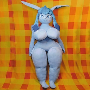 May include: A large, plush doll in a light blue color, featuring a stylized character design with large eyes, long ears, and a distinctive headpiece. The doll has a rounded body shape and is set against a yellow and orange checkered background.