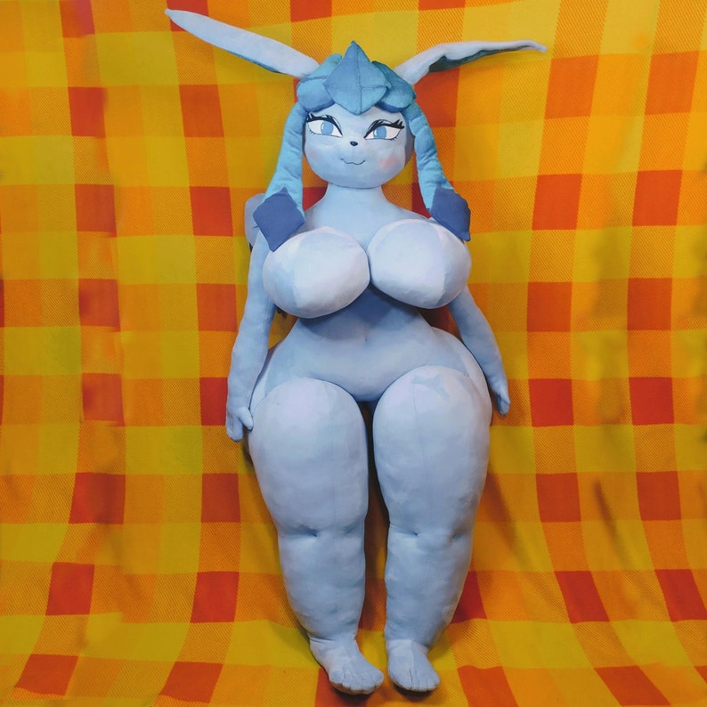 May include: A large, plush doll in a light blue color, featuring a stylized character design with large eyes, long ears, and a distinctive headpiece. The doll has a rounded body shape and is set against a yellow and orange checkered background.