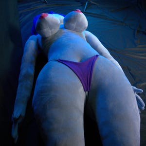 May include: A plush, light gray figure in a reclining pose, wearing a purple bikini bottom. The figure has large breasts with pink nipples and blue hair. The background is a dark, textured surface.