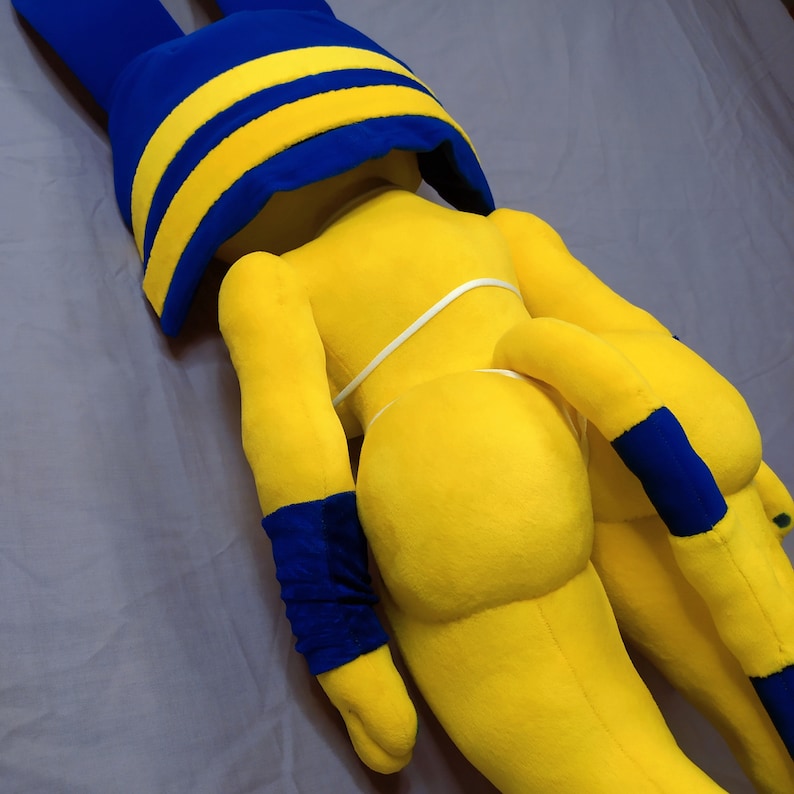 Puede incluir: Un juguete de peluche amarillo con una cubierta de cabeza azul y amarilla. El juguete tiene una cola larga y est&aacute; acostado sobre una superficie gris.