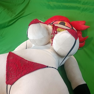 Puede incluir: Una mu&ntilde;eca de peluche grande con un cuerpo blanco, que lleva un bikini de encaje rojo. La mu&ntilde;eca tiene pechos grandes y redondos y una cabeza roja con una cara de dibujos animados. El fondo es verde liso.