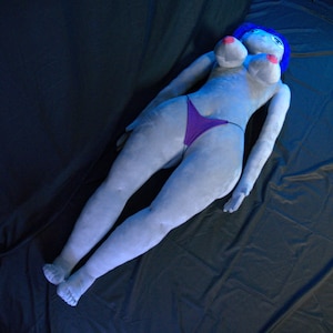 May include: A plush doll in the shape of a reclining figure, with blue hair, light skin, and a purple thong. The doll has large breasts with pink nipples. The doll is lying on a dark surface.