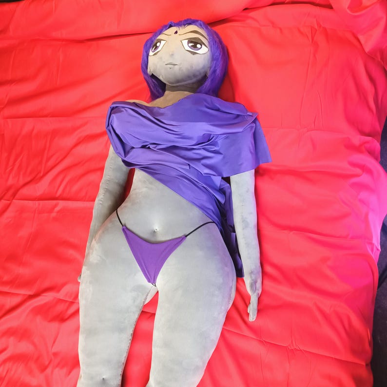 May include: A plush doll of a cartoon character, featuring a gray complexion, purple hair, and a purple outfit. The doll is lying on a red surface, with the character's face and body details clearly visible. The doll is wearing a purple top and a purple thong.