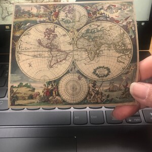 May include: A vintage-style world map print featuring detailed illustrations of continents and oceans. The map is in a square format, with a beige background and intricate drawings of figures and ships. The print is held over a laptop keyboard.