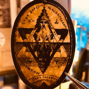 May include: Oval sticker with a sepia-toned illustration of a bearded figure within a star, surrounded by text. The figure wears a crown and holds a measuring tool. The sticker has a black border and is held by tweezers.