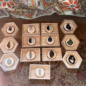 May include: A collection of 11 wooden coasters with various natural stone and crystal accents. Each coaster features a different stone or crystal, including amethyst, clear quartz, black obsidian, and green aventurine. The coasters are arranged in a grid pattern on a light brown surface.
