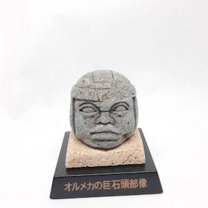 May include: A gray stone sculpture of a head with a flat top and a serious expression. The sculpture is on a small, rectangular base with the text "Olmeca's Stone Head Replica" in Japanese.