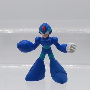 Könnte beinhalten: Eine blaue und türkisfarbene Plastik-Actionfigur eines Roboters mit rotem Visier. Der Roboter hat eine weiße Hand und ist in einer Pose mit einem ausgestreckten Arm.