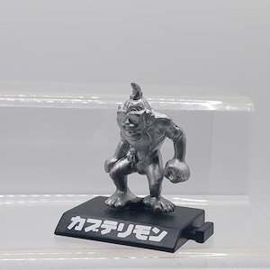 May include: A silver figurine of a creature with a pointed head, wearing sunglasses and holding two weights. The figurine is standing on a black base with the Japanese text "カステリモン" written on it.