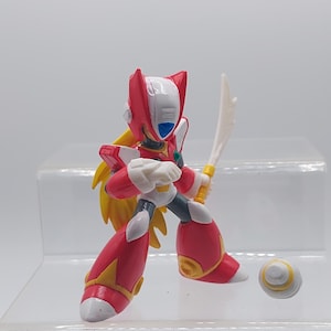 May include: A red and white action figure of a robot character holding a sword. The robot has a yellow and black helmet and is standing on a clear plastic base. A yellow and white ball is in the foreground.