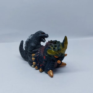 May include: A black and gold Godzilla kaiju figure with a smaller black Godzilla figure on top. The larger figure has red and gold accents and a spiky back.