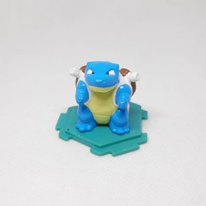 May include: A blue and yellow Pokemon figurine of Squirtle standing on a green base. The figurine is wearing a white shell and has a brown shell on its back.