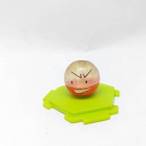 Può includere: Un piccolo giocattolo di plastica arancione e bianco con un'espressione arrabbiata. Il giocattolo è seduto su una base esagonale verde.