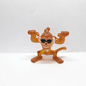 May include: A small, brown plastic monkey figurine wearing sunglasses. The monkey is standing with its arms raised and its legs spread apart. It has a yellow star on its chest.