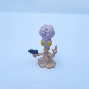 May include: A small, alien figurine with a large, purple brain-like head. The figure has yellow eyes, a tan body, and is holding a black ray gun. One finger is raised in a gesture. The figurine is on a light blue background.
