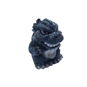 Puede incluir: Una pequeña figura de plástico gris oscuro de un monstruo parecido a Godzilla con dientes afilados y una expresión amenazante.