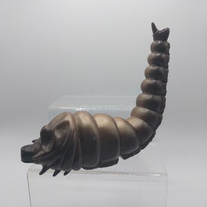 May include: A brown, segmented, insect-like sculpture with a curved body and a detailed head. The sculpture has a textured surface and is displayed on a clear, tiered platform. The overall design suggests a creature or insect.
