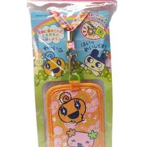 Puede incluir: Lanyard naranja con un personaje de dibujos animados rosa y blanco en la parte delantera. El lanyard tiene un porta-tarjetas de identificación a juego naranja y rosa con un personaje de dibujos animados rosa y blanco en la parte delantera. El texto en el porta-tarjetas dice "TMG".