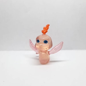 Puede incluir: Una pequeña figura de dragón de plástico rosa translúcido con alas naranjas y ojos azules. El dragón tiene una pequeña cresta naranja en la cabeza.