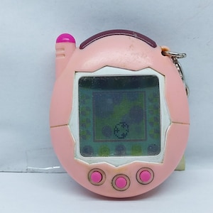 May include: A pink and white digital pet toy with a screen displaying a green and white image. The toy has three pink buttons and a pink antenna.