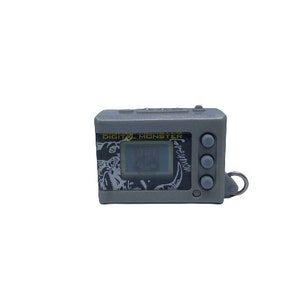 May include: A gray plastic digital pet toy with a black and white screen displaying the text "Greymon". The toy has three buttons on the right side and the text "Digital Monster" on the top.