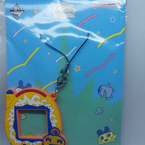 May include: A yellow and orange character keychain with a blue and white screen. The keychain has a black string and a silver clip. The packaging has a blue background with colorful lines and stars. The text on the packaging says "BANDAI NAMCO" and "たまごっち!"