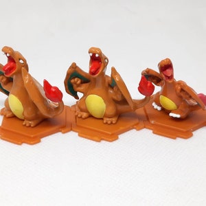 May include: Three orange and yellow plastic figurines of a Pokemon character, Charizard, with red flames and green wings. The figurines are posed in different positions and are standing on hexagonal bases.