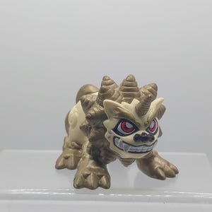 Peut inclure: Une petite figurine brune et blanche d'une créature mythique avec des cornes et des yeux rouges. La créature a une expression féroce et semble grogner.