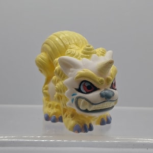 Peut inclure: Une figurine jaune et blanche d'une créature mythique à l'expression féroce. La créature a une crinière longue et fluide et une corne sur la tête. Elle a des yeux rouges et un large sourire aux dents acérées.